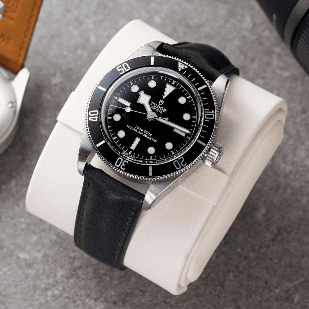 Watch with black leather strap and black dial on a white cushion against a gray background