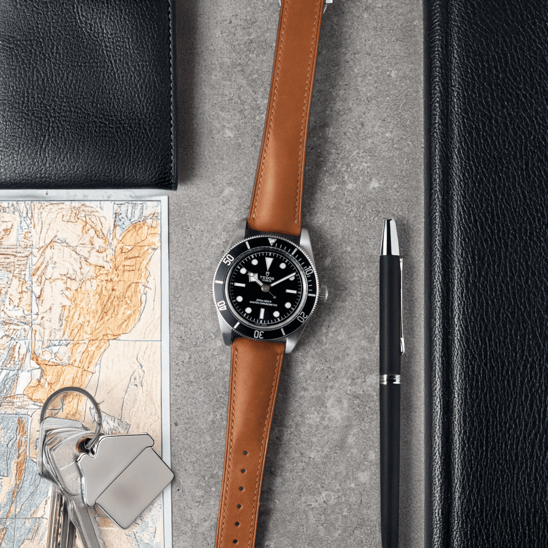 Watch with brown leather strap on a gray surface with map, keys, and pen.