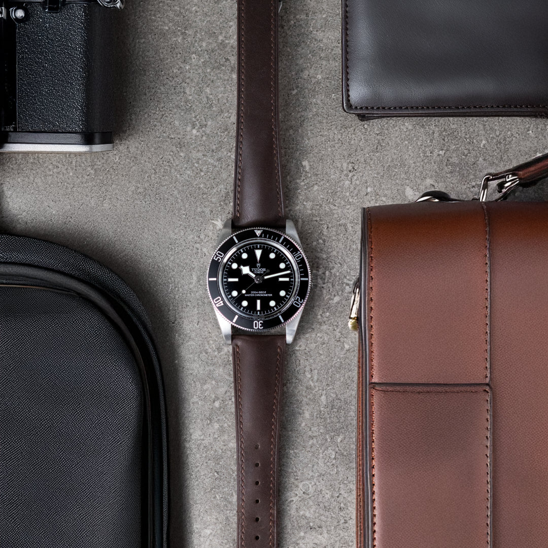 Watch with brown strap on a textured surface with bags and a wallet