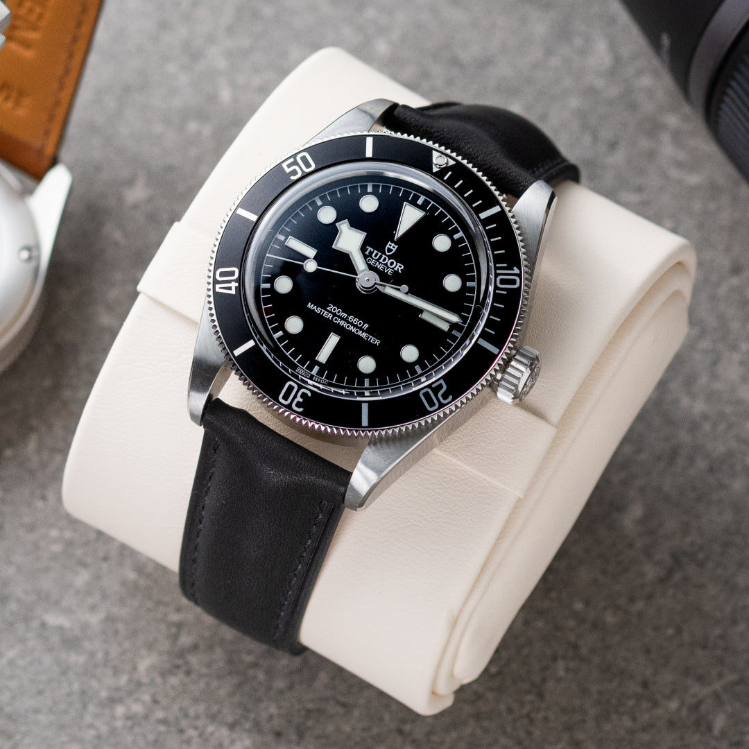 Watch with black leather strap and black dial on a white cushion against a gray background