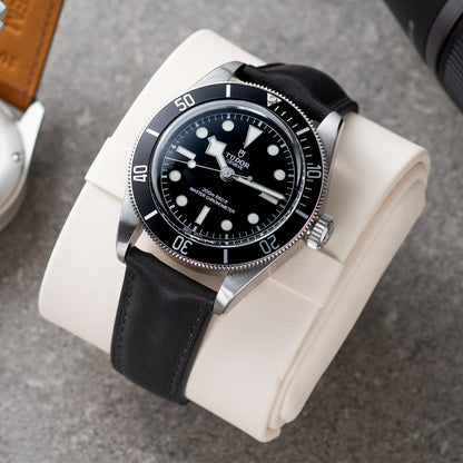 Watch with black leather strap and black dial on a white cushion against a gray background
